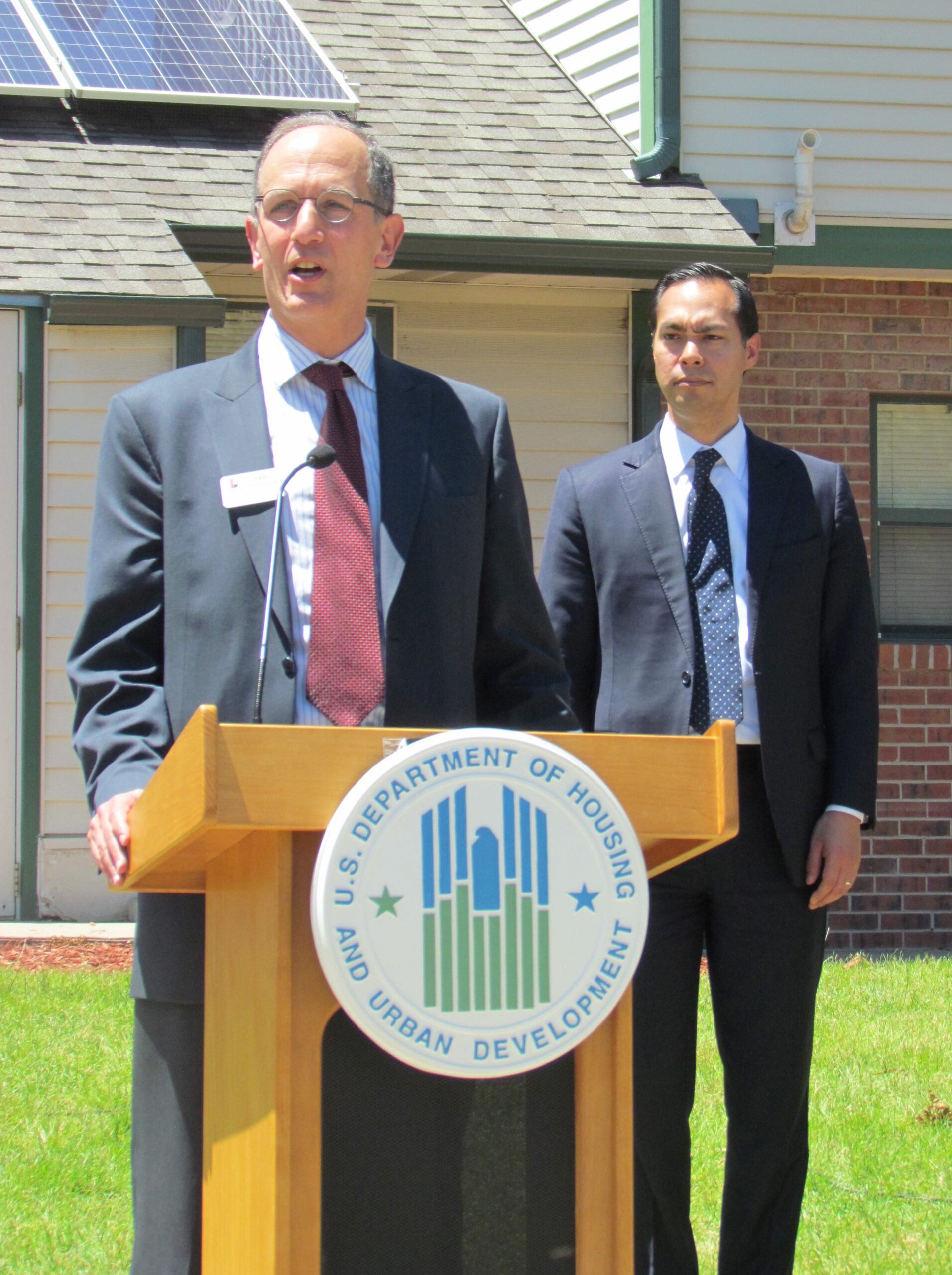 Jerry Tinianow standing at a podium speaking with then-Secretary Julian Castro of the U.S. Department of Housing and Urban Development standing behind him